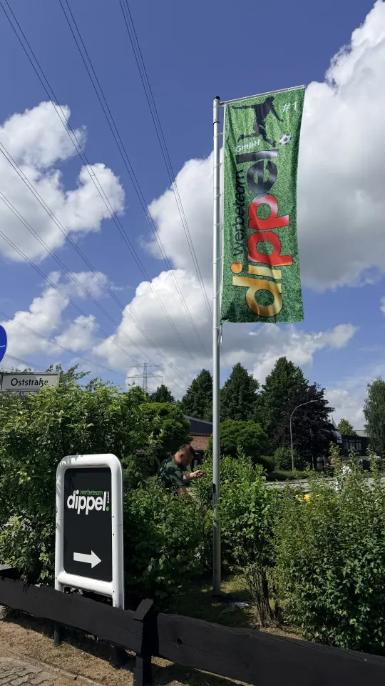 Hochformat-Flagge von Werbeteam Dippel am Fahnenmast an der Abzweigung zur Oststraße in Norderstedt – rund 50 m vor unserem Firmensitz